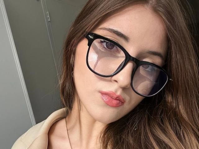 Maisie Vandenberghe mit großen Brillen, sinnlich geschürzten rosa Lippen und braunen, welligen Haaren, trägt ein beiges Oberteil und blickt mit ernstem Gesichtsausdruck in die Kamera..