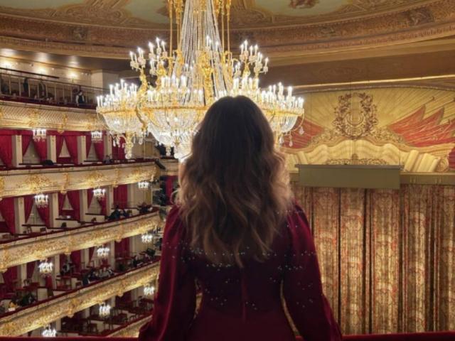 Maisie Vandenberghe, con el pelo largo y ondulado de color marrón, vestida con un vestido rojo con lentejuelas doradas, está de pie frente a un gran candelabro dorado y blanco, muy brillante, en una habitación ornamentada..