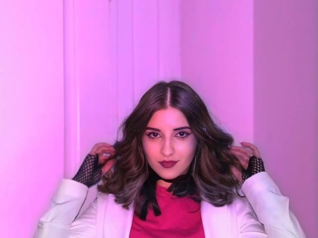 Maisie Vandenberghe, con cabello castaño ondulado, vestida con un blazer blanco, un cuello alto rojo y guantes de malla negra, tiene un rostro adorable y labios carnosos, en una habitación iluminada con luz rosa..
