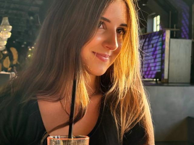 Maisie Vandenberghe, con lunghi capelli castani, sorridente, con il sole sul viso, indossa una maglietta nera e tiene in mano una bevanda con una cannuccia nera. L'immagine è caratterizzata da una luce calda e soffusa, un'atmosfera casual e rilassata, e trasmette un senso di bellezza..