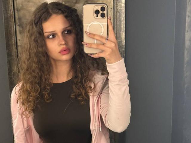 CurlyElina met krullend bruin haar, een roze vest, een zwarte top, die een telefoon vasthoudt, zijwaarts kijkt, roze lippen, een serieuze uitdrukking, een blauw-grijze muur, een zichtbare telefooncamera met een ring..