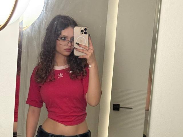 CurlyElina met krullend bruin haar, een bril, een rood Adidas shirt, waarbij haar buik zichtbaar is, die een iPhone vasthoudt voor een spiegelzelfportret, staand in een badkamer, met donkere broek, reflectie zichtbaar..