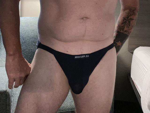 Braadley69's bare torso and black, tight, boy-style underwear, showing a bulge, with tattoos on the right arm, standing in a dimly lit room.