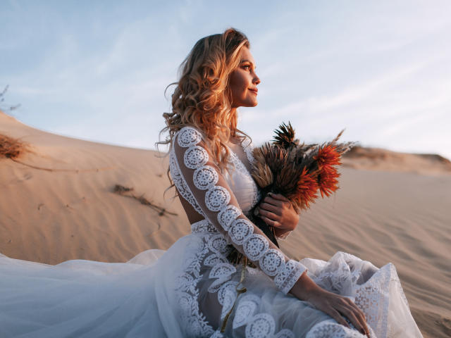 EolantaMarina with curly blonde hair, wearing a white lace dress, holding a bouquet of orange and brown feathers, sitting in sandy dunes, sunlight on her profile, serene expression.