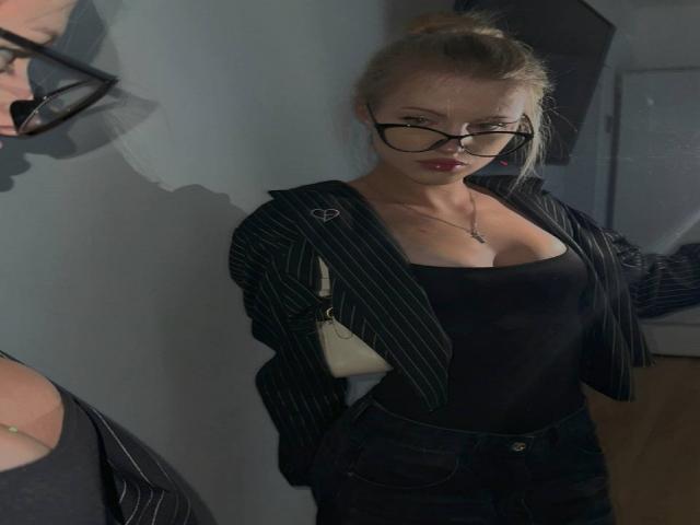 JulissaUrso met blond haar in een knot, een zwart colbert met strepen, een zwart topje dat haar borsten laat zien, en donkere jeans, draagt een bril en een ketting, en staat in een slecht verlichte kamer..