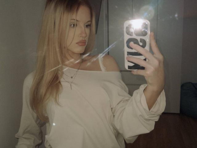 JulissaUrso, loira, com cabelo comprido, usando uma blusa branca com um ombro descoberto, segurando o celular em um selfie no espelho, com reflexo do flash, mostrando a parte superior do corpo, seios pequenos, chão de madeira, leve efeito de lente..