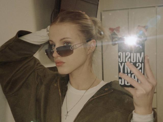 Julissa Urso, met blond haar in een knot, draagt een zonnebril van het type "aviator", een groene jas, een wit shirt en rode lippenstift, en maakt een selfie met flits. Haar hand bevindt zich op haar hoofd..