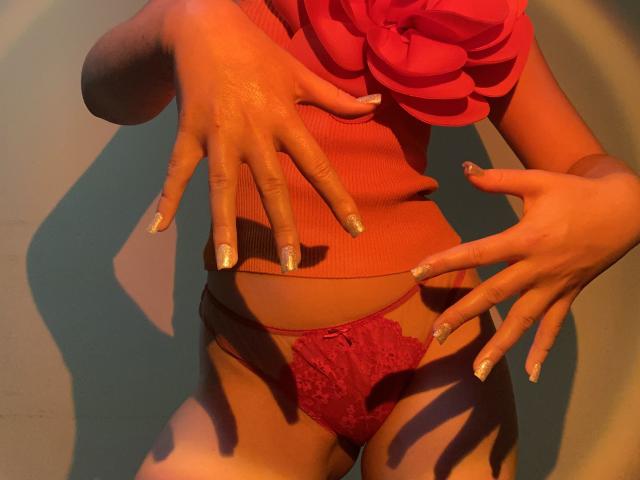 La lingerie rossa in pizzo di FlyFreedom, la maglietta arancione a coste con fiore, le mani con le unghie scintillanti, che mette in mostra la sua abbronzatura e il suo sedere sexy e formoso. L'illuminazione calda e ombreggiata esalta la sua figura..