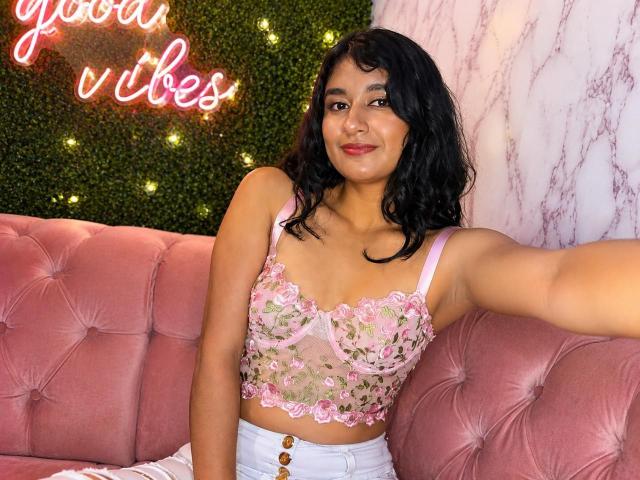 Adara Rivat, con piel de tono marrón medio, cabello negro ondulado, un corpiño de encaje rosa que deja ver sus pechos, pantalones blancos de cintura alta, un sofá rosa con adornos, un letrero de neón, sonriendo, selfie..