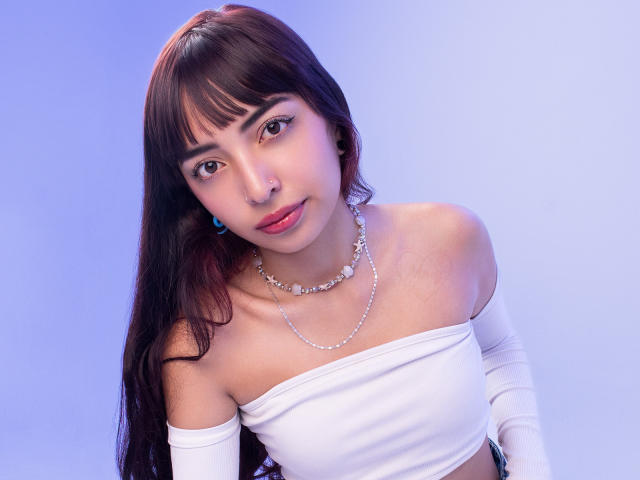 IrisVelvet, con cabello castaño oscuro, vestida con una blusa blanca con un hombro descubierto y un collar de perlas, tiene los pechos ligeramente expuestos, mirando hacia abajo con una sonrisa sutil. Fondo azul claro..