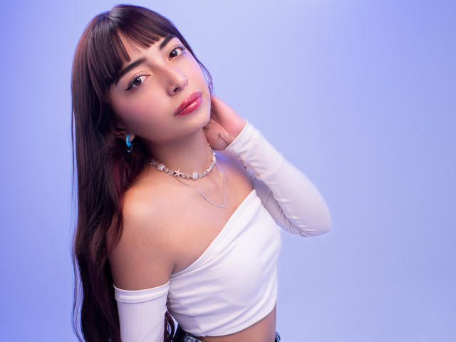 IrisVelvet ha lunghi capelli castani, indossa una maglietta bianca con una spalla scoperta, orecchini blu e una collana floreale, con una mano al collo, e guarda verso la fotocamera..