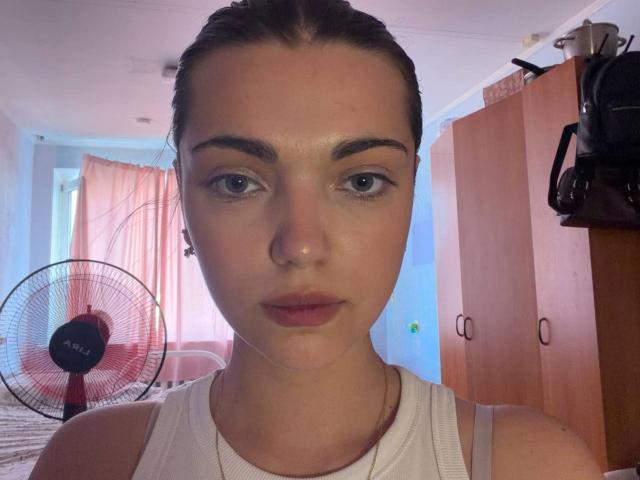 PatriciaKusick heeft een lichte huid, blauwe ogen en bruin haar in een paardenstaart, draagt een witte tanktop en kijkt recht in de camera met een neutrale uitdrukking..
