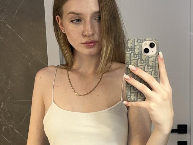 Ginnia mit hellbraunen Haaren, rosafarbenen Lippen und einer schlanken Figur, trägt ein weißes Tanktop und hält ein Handy mit einer gemusterten Hülle. Sie zeigt ihre Brüste und eine goldene Kette..