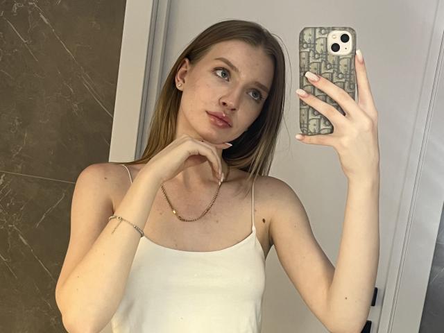 Ginnia with fair skin and long blonde hair, wearing a white tank top, holding a phone with a patterned case, looking in the mirror, touching her chin.