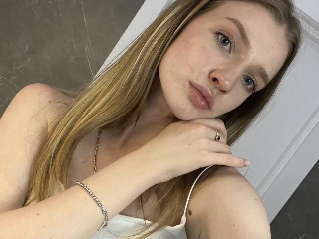 Ginnia, met blond haar, lichte huid en blauwe ogen, draagt een witte top en heeft haar kin in haar hand, met een subtiele glimlach, en een zilveren armband om haar pols..