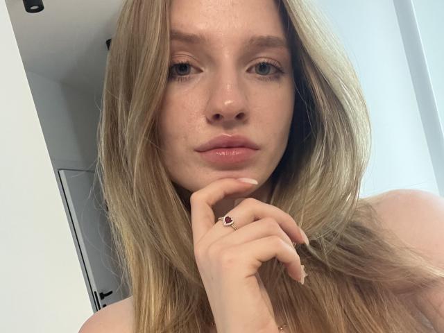 Het gezicht van Ginnia, met blond haar, sproeten en glanzende, roze lippen, die haar kin aanraakt met een ring aan haar vinger, waarbij haar borsten lichtjes in de voorgrond te zien zijn..