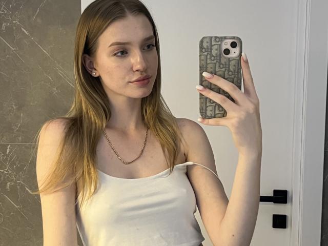 Ginnia, avec ses cheveux blonds, portant un débardeur blanc, tenant un téléphone avec une coque à motifs, prenant un selfie devant un miroir, montrant ses seins, et affichant un léger sourire..