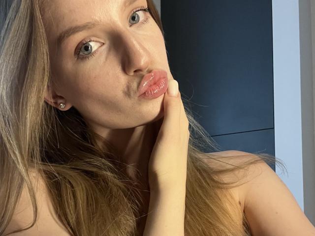Ginnia with pouty pink lips, blue eyes, and blonde hair, touching her chin with her hand, showing off her bare shoulders. She's wearing small earrings.