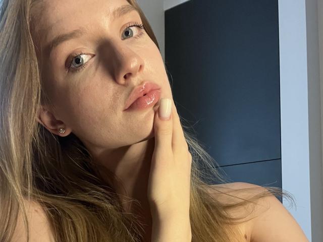 Ginnias Gesicht, mit blonden Haaren, blauen Augen und glänzenden rosafarbenen Lippen, berührt ihr Kinn. Sie trägt einen kleinen Ohrring, und ihre Hand berührt sanft ihr Gesicht..