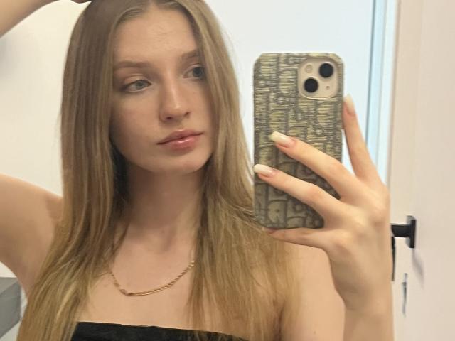 Ginnia, con cabello rubio largo, sosteniendo un teléfono en su mano derecha, mostrando su rostro y un top negro con tirantes, llevando un collar dorado, en un selfie frente a un espejo..