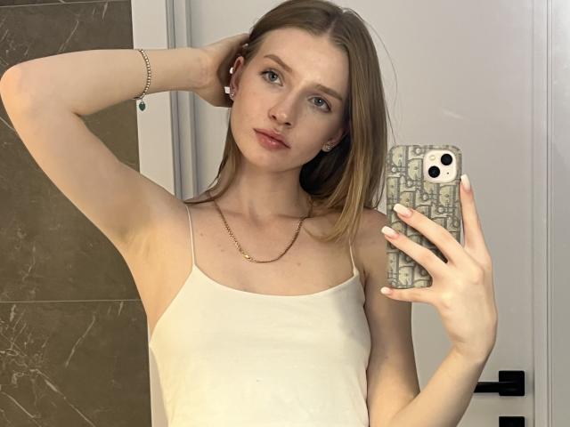 Ginnia, con la pelle chiara e gli occhi azzurri, indossa una canottiera bianca, tiene in mano un telefono con una custodia marmorea, ha un braccio alzato e guarda la fotocamera, indossa una piccola collana dorata..