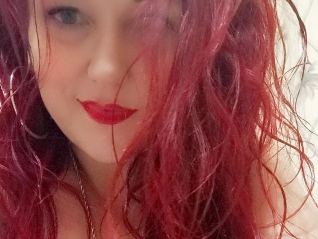 HIMandHER mit roten, gewellten, feucht aussehenden Haaren, die die Hälfte ihres Gesichts bedecken, heller Haut, rotem Lippenstift und einem subtilen Lächeln, wobei ein Nasenring und eine Kette zu sehen sind..