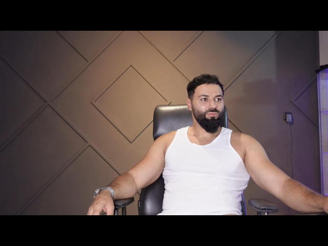 MattMasters, un uomo con la barba e una corporatura muscolosa, che indossa una canotta bianca, seduto su una sedia nera con le braccia aperte, con un'espressione leggermente infastidita..