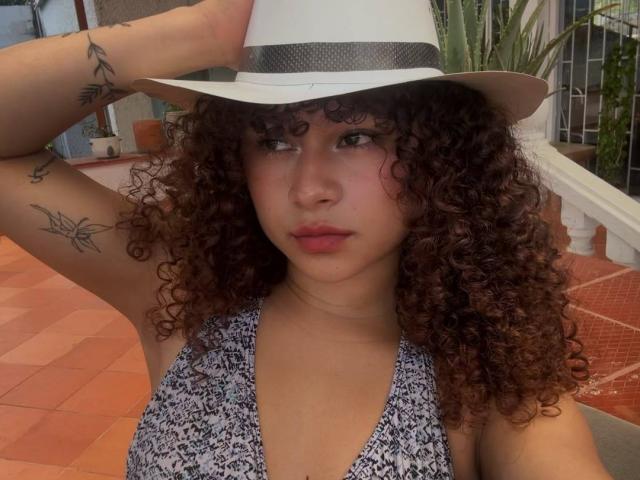 Sofia, met krullend bruin haar, draagt een witte hoed, een blauw-witte top en heeft tatoeages op haar arm. Ze kijkt serieus in de camera..