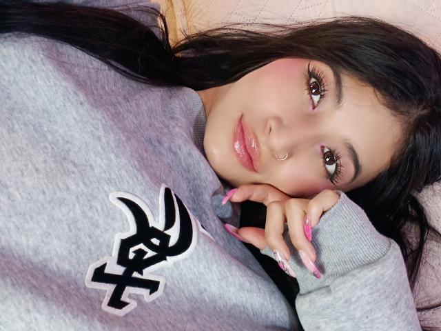 Naia Sanders, con cabello negro largo, labios rosados y brillantes, un aro en la nariz, ojos marrones y uñas rosadas, está acostada sobre un cojín, vistiendo una sudadera gris con un logo negro..