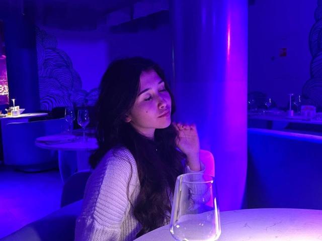 YingKeeter, com cabelos longos e escuros, vestindo um suéter branco de tricô, sentada à mesa com um copo, olhando ligeiramente para o lado sob a iluminação roxo-azul de uma boate..