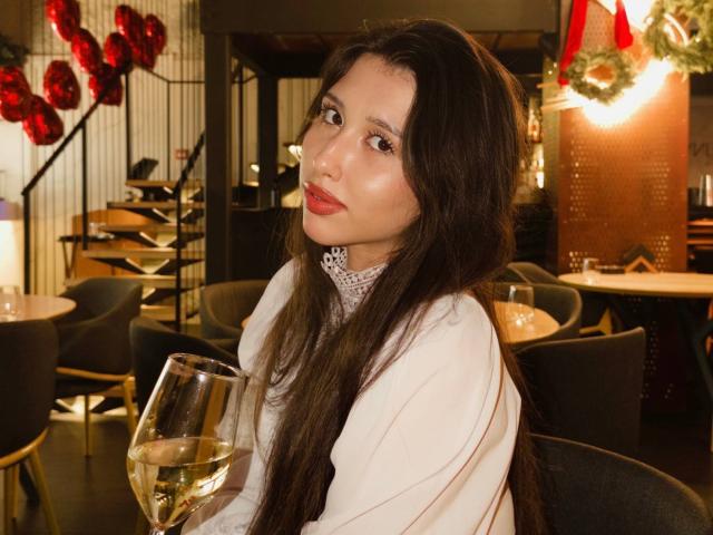 YingKeeter, con cabello castaño largo, lápiz labial rojo, un cuello de encaje blanco, sosteniendo una copa de vino blanco, sentada en un restaurante tenuemente iluminado con decoraciones rojas y luces de Navidad..