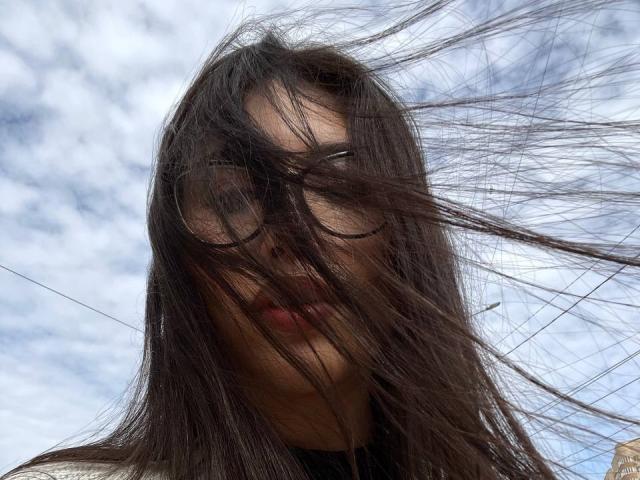 El largo cabello castaño de YingKeeter ondea salvajemente con el viento, cubriendo sus ojos y parcialmente su rostro, con sus labios ligeramente visibles. Lleva gafas de sol reflectantes..
