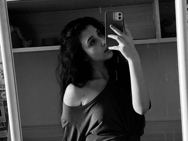 YingKeeter, com cabelo escuro e ondulado, camisa com um ombro de fora e a língua para fora, tirando uma selfie com um iPhone, mostrando um pouco o colo e traços femininos suaves..