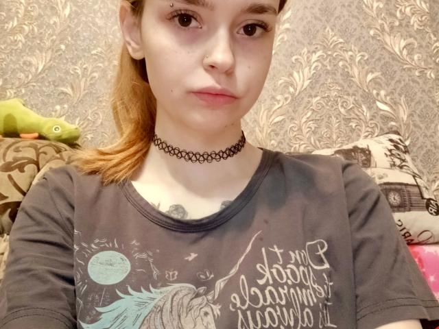 Merri Serafin, mit heller Haut, braunen Augen und einer schwarzen Halskette, trägt ein graues T-Shirt, hat einen blonden Pferdeschwanz und eine kleine Nasenring, und sitzt in einem gemütlichen Raum..