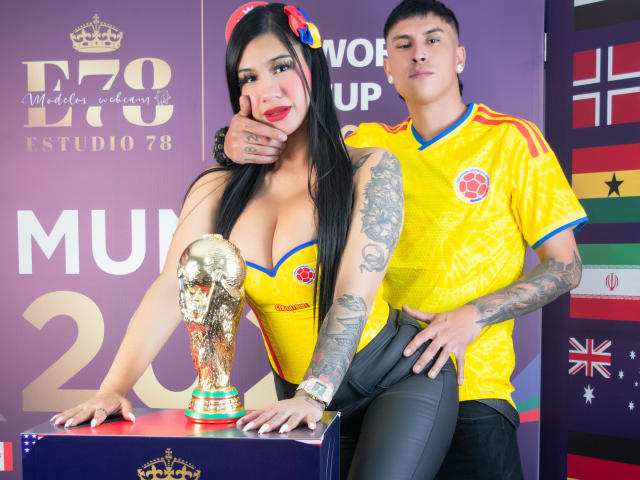 Kathy e Zac, com seios grandes, vestindo uma camisa esportiva amarela e calças de couro pretas, estão ao lado de Zac, que tem tatuagens, e ele segura um troféu dourado. Kathy tem cabelos longos e pretos, e Zac parece confiante..