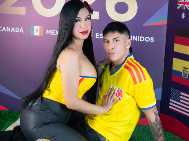 KathyAndZac met lang zwart haar, rode lippenstift en grote borsten in een gele top, die Zac's borst aanraakt terwijl hij een geel voetbalshirt van Mexico draagt. Zac heeft tatoeages en een kort kapsel..