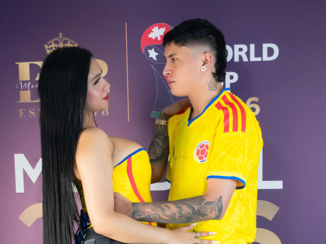 Kathy y Zac, con cabello negro largo, busto grande y vistiendo una camiseta de fútbol amarilla, y Zac con cabello negro corto, tatuajes y también vistiendo una camiseta de fútbol amarilla, están de pie cerca, mirándose el uno al otro..