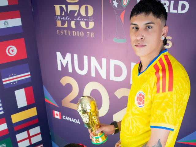 KathyAndZac, com cabelo preto curto, pele de tom médio e alargadores de lóbulos, vestindo uma camisa amarela de futebol, segurando um troféu dourado da Copa do Mundo da FIFA, sorrindo para a câmera..