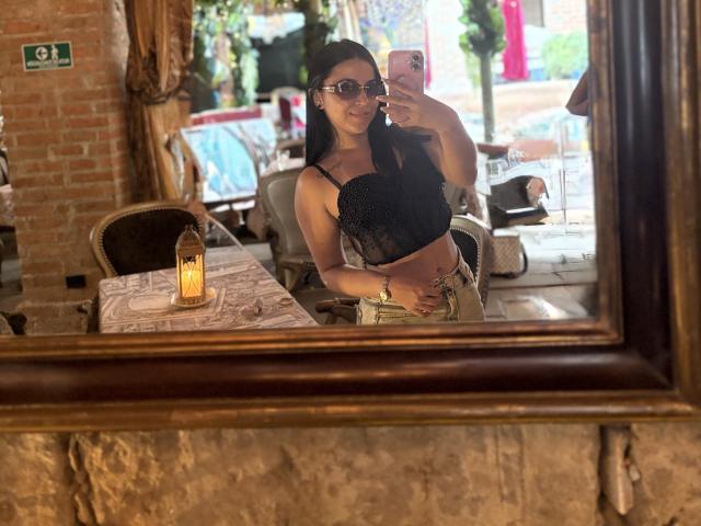 HikariChloe, com cabelo escuro, óculos, blusa de renda preta e calças bege, tirando uma selfie no espelho em um café aconchegante e iluminado. Seios visíveis, mão na barriga..