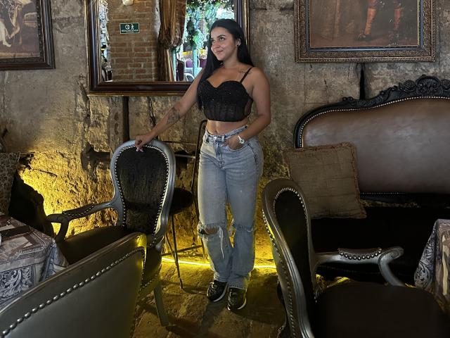 HikariChloe com uma blusa preta de renda, mostrando os seios, vestindo jeans rasgados, em pé em um quarto com cadeiras ornamentadas, sorrindo, com o braço apoiado na cadeira, tatuagem no antebraço esquerdo..