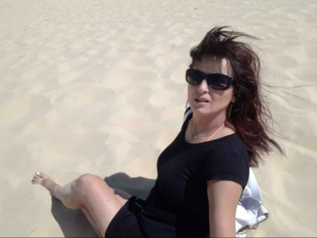 YummyTitss con gafas de sol oscuras, camisa negra y pantalones cortos negros, sentada en arena mojada, con las piernas cruzadas, el cabello castaño ondeando, mirando hacia arriba, descalza, con una toalla blanca debajo..