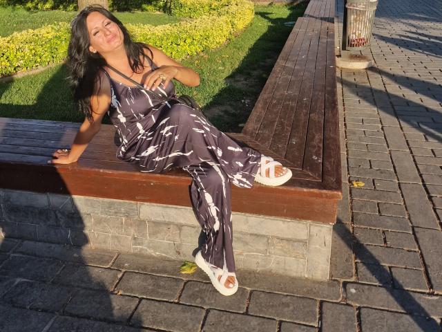 YummyTitss with dark hair, eyes closed, smiling, sitting on wooden bench in black patterned dress, white sandals, hand on chest, sunlit, brick path, grass in background.