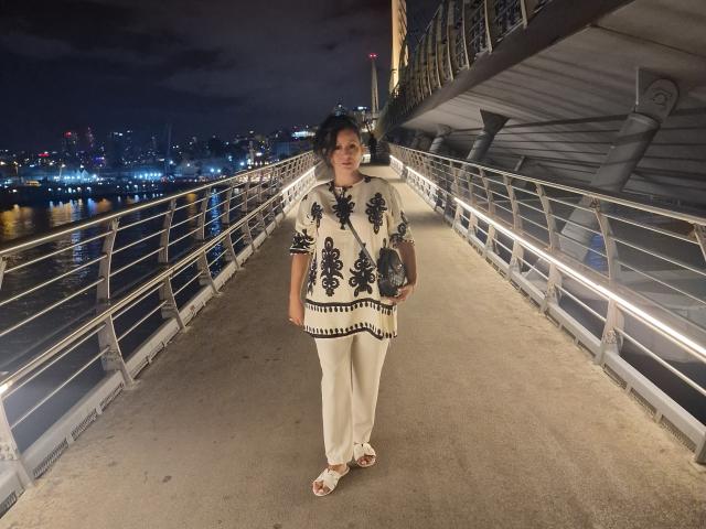 YummyTitss stands on a bridge at night, wearing a white blouse with black floral patterns, beige pants, and white sandals, holding a gray purse, looking forward.