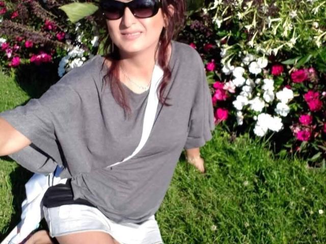 YummyTitss with brown hair, sunglasses, gray shirt, white shorts, and black strap, sitting on green grass, smiling. Boobs not visible, ass not visible, pussy not visible.