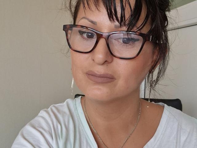 YummyTitss, avec des lunettes marron, des seins de taille moyenne, et un t-shirt blanc, a une expression sérieuse, une peau claire, des cheveux bruns, et porte un collier en argent..