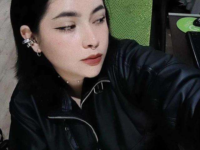 Phylavell mit heller Haut, schwarzen Haaren und auffälligem Eyeliner, trägt eine schwarze Lederjacke, hat eine Nasenring und mehrere Ohrringe, blickt nach unten mit leicht geöffneten, roten Lippen..