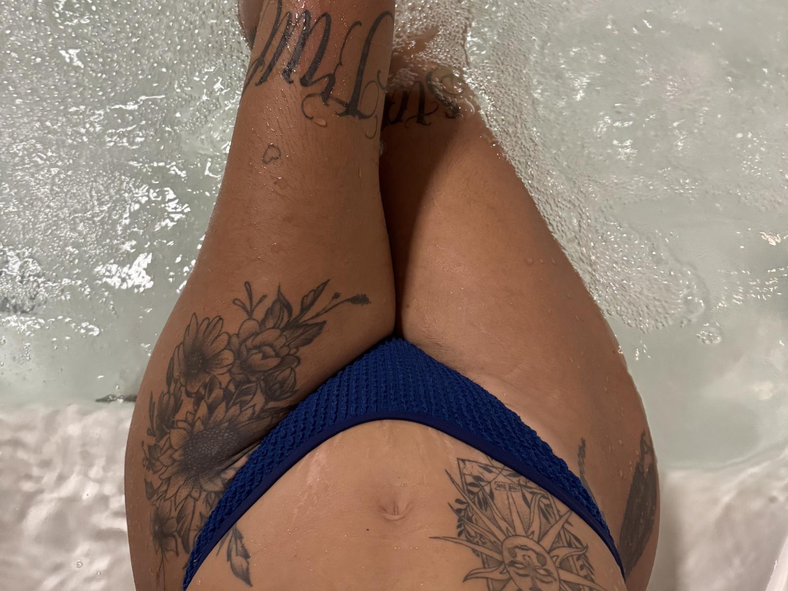 RamonaGs Beine in einem blauen Bikini-Unterteil, mit Blumen und Schrift tätowiert, nass vom Wasser, sichtbare Tattoos an Oberschenkeln und Waden, Wassertropfen auf der Haut, Beine angewinkelt..