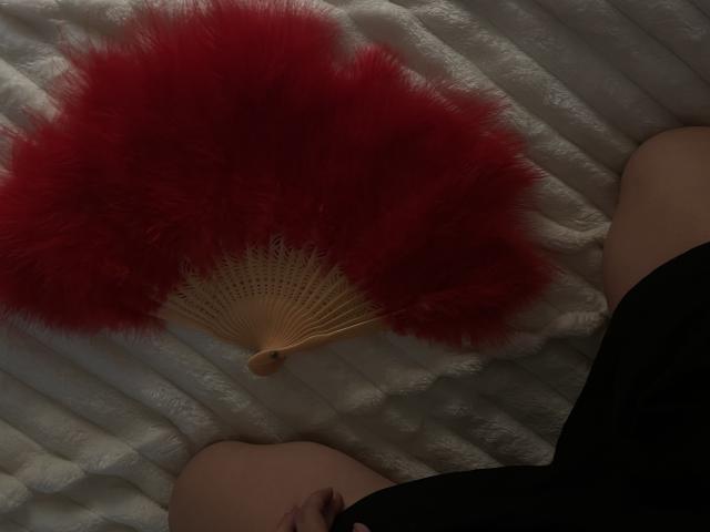 AlexXas's red fluffy hand fan rests on their bare thighs, with black underwear, in a dimly lit, textured white surface. Their pale skin is visible.