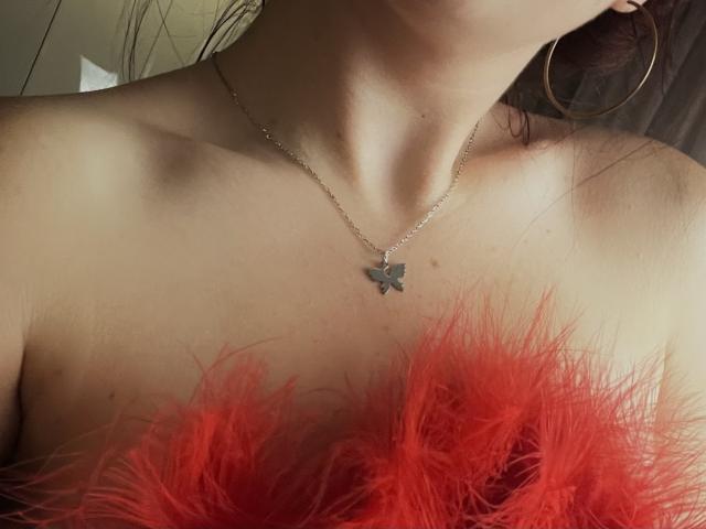 AlexXas's bare shoulders with a red feathered top, wearing a butterfly necklace and large hoop earrings, her collarbone and neck visible. Her skin is light.