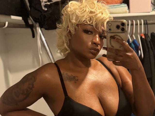 SunnyMochaQueen con capelli biondi ricci, pelle marrone scuro, reggiseno nero, tatuaggi sul braccio sinistro, che tiene un telefono, selfie allo specchio, seno grande, espressione seria, unghie scure, custodia del telefono con un anello..